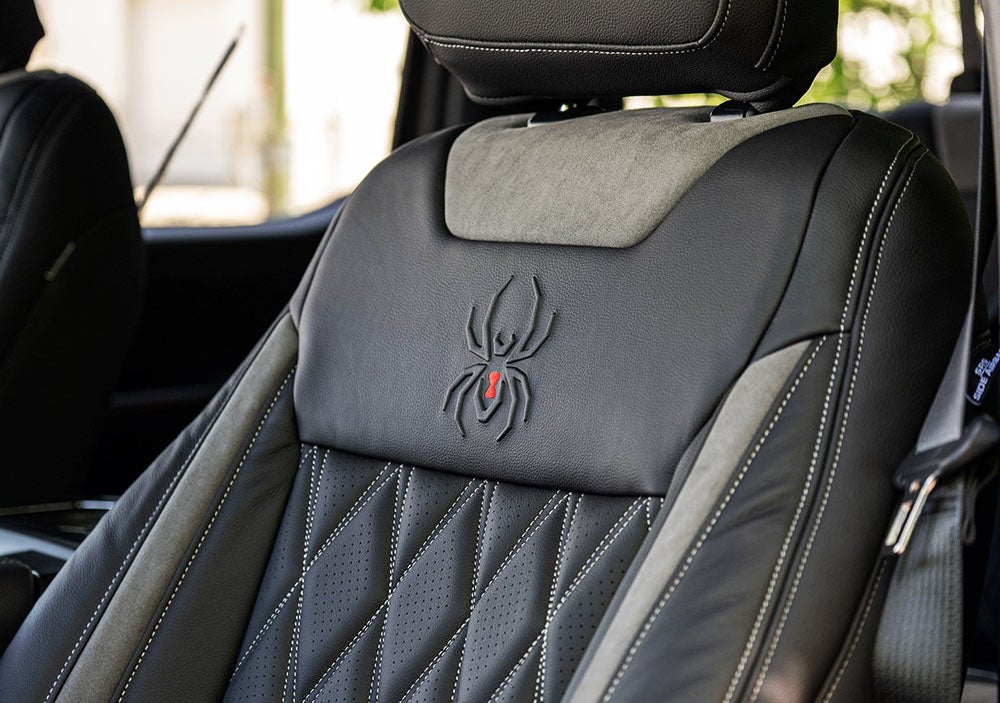 Close-up of a car's front seat with black leather upholstery, featuring a stitched black widow emblem with a red mark on the seat backrest.